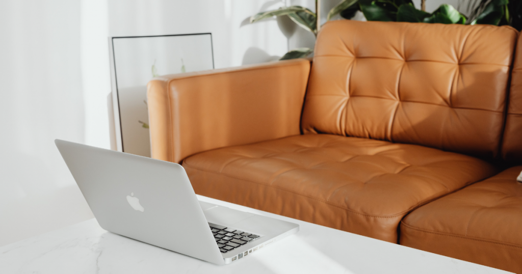 A tall coffee table with a laptop on it besides a brown leather sofa