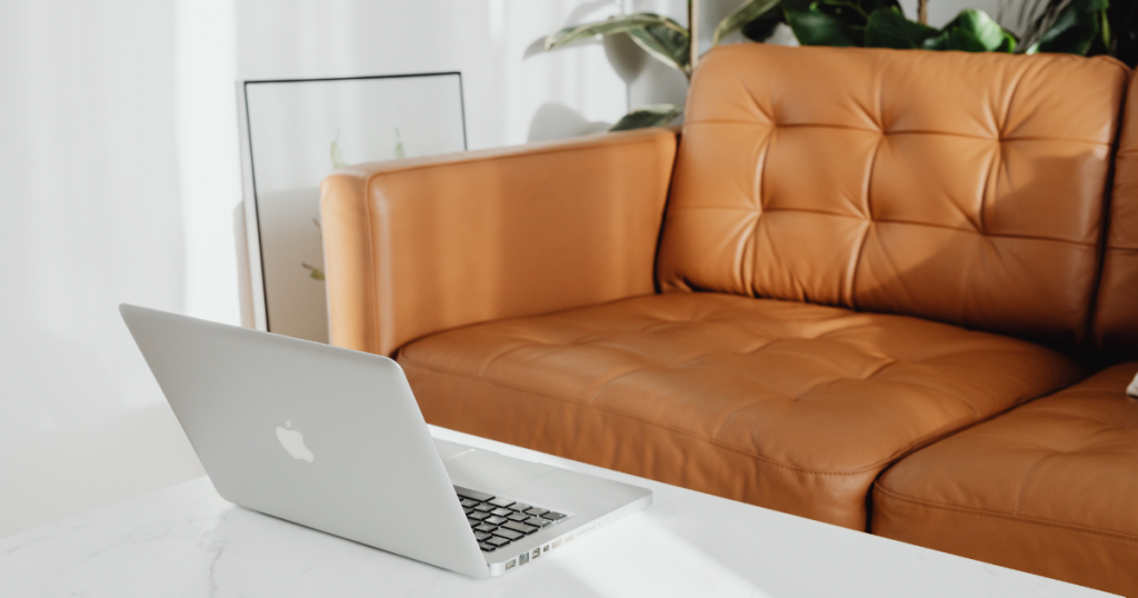 A tall coffee table  with a laptop on it besides a brown leather sofa
